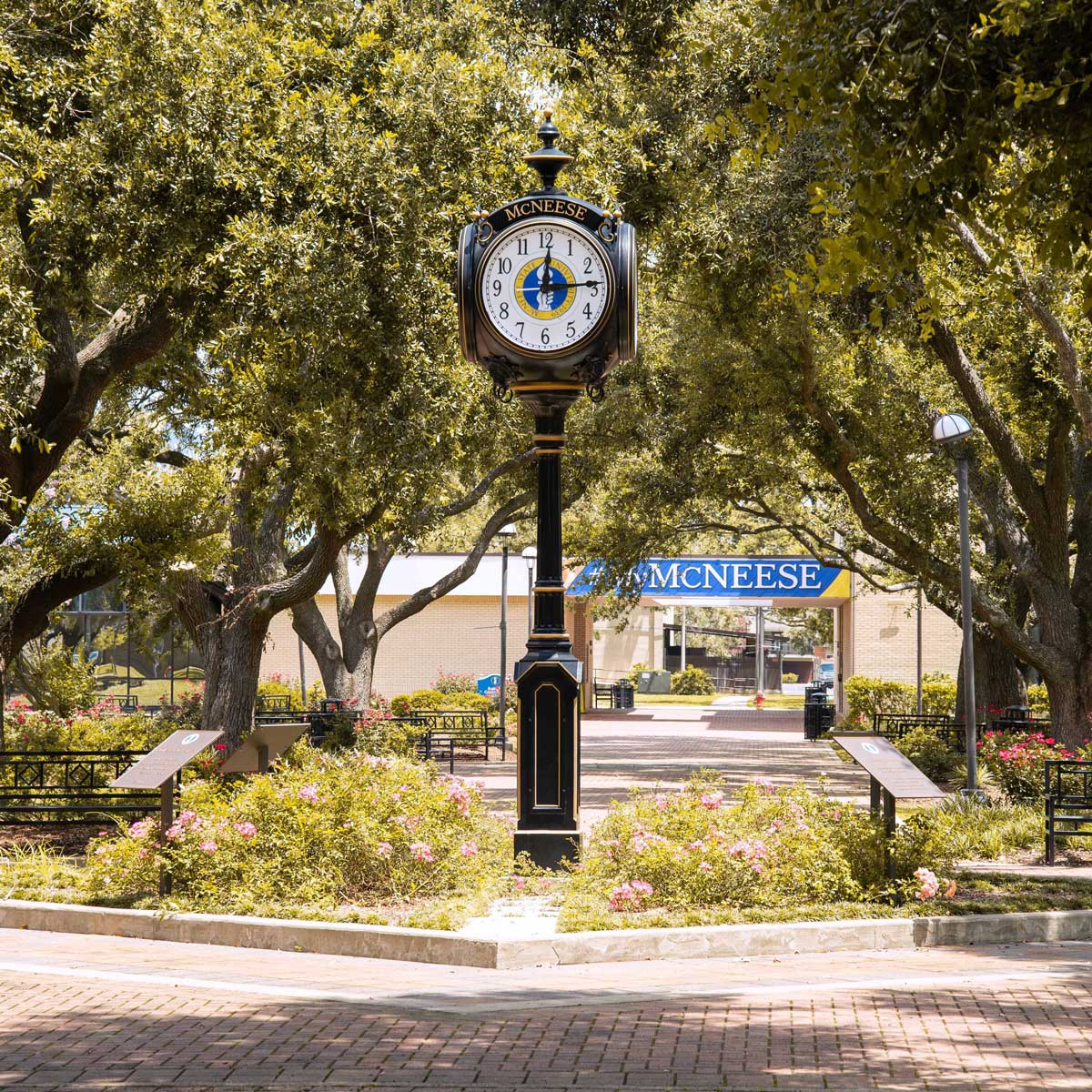 Black clock tower with McNeese sign in background, surrounded by trees and flowers on a sunny day.