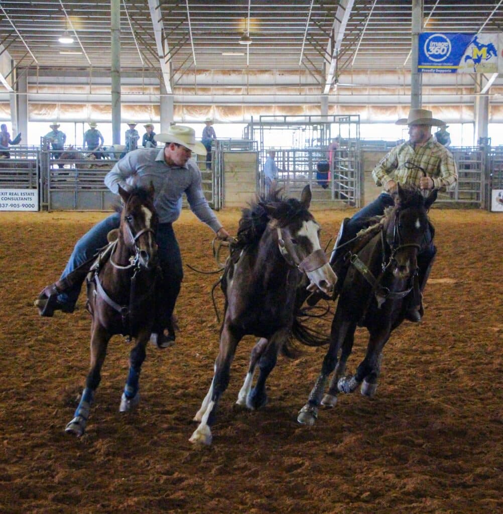 Two cowboys performing at a rodeo inside an arena, riding horses at full speed, surrounded by metal fences.