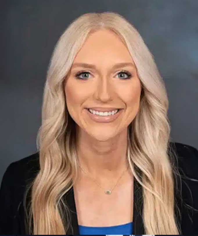 Smiling woman with long blonde hair wearing a black blazer and blue top, neutral background.