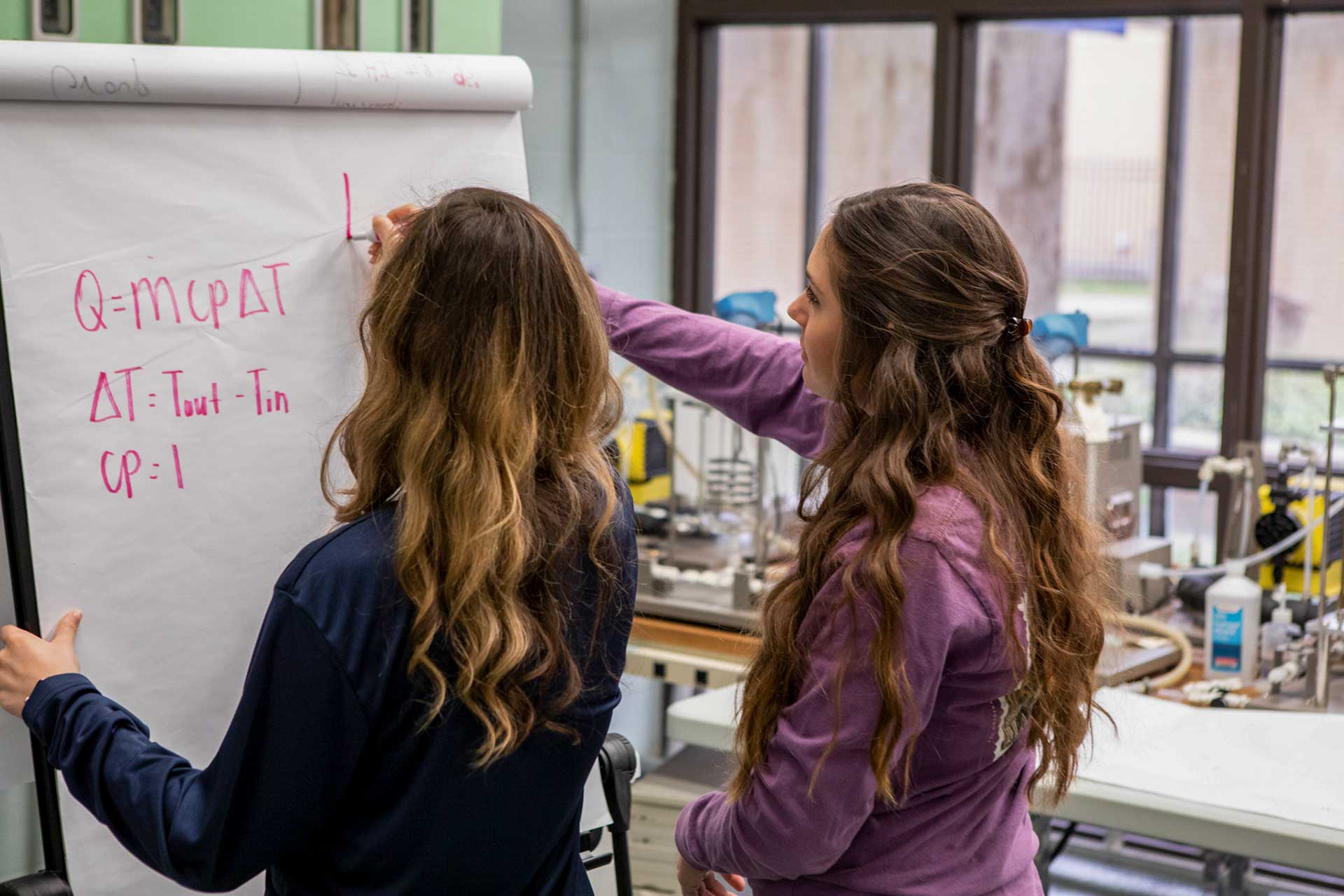 Two students solving a thermodynamics equation on a whiteboard in a lab setting.