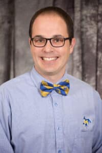 Smiling man in glasses wearing a blue shirt with logo and yellow-blue bow tie.