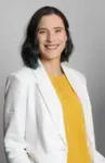 Smiling person in a white blazer and yellow top against a gray background.